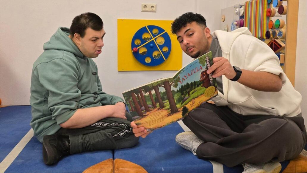Twee mannen zitten op een kleed en de een leest de ander een boek voor